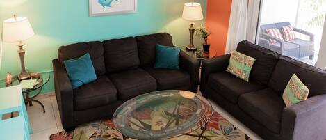 A living room with two dark sofas, a glass coffee table, and a colorful floral rug. There are two lamps on side tables, a framed picture on the wall, and a view of the outdoors through a window.