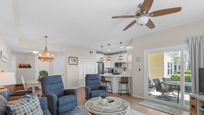 A spacious living room with a ceiling fan, a dining area with a kitchen in the background, and a sliding glass door leading to an outdoor patio. - Beautifully Updated 2BR Beachwalk (Destin)