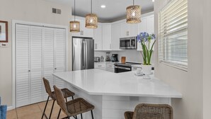 A modern kitchen with white cabinets, a large island, wicker chairs, and pendant lights. - Beautifully Updated 2BR Beachwalk (Destin)