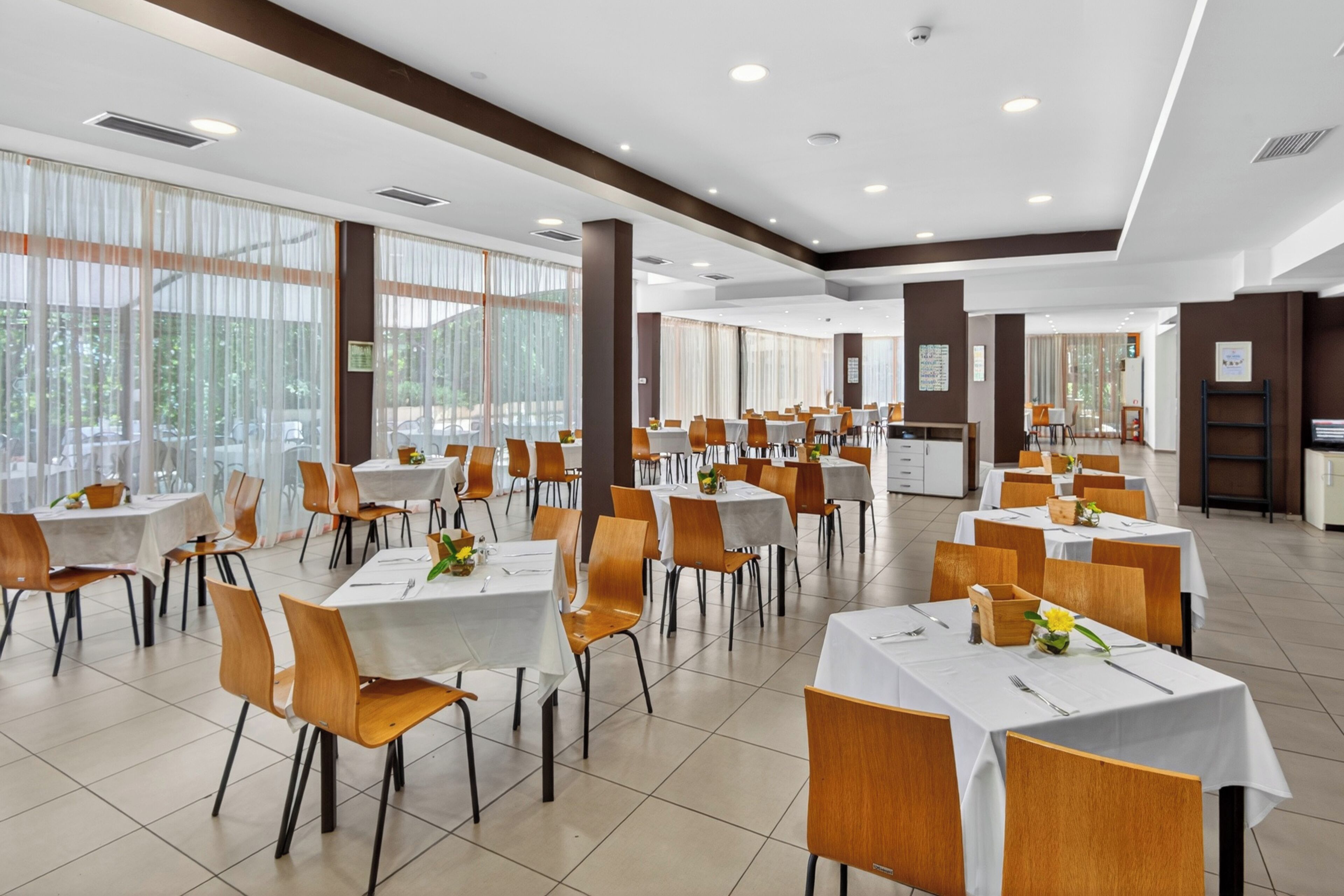 A spacious dining area with multiple tables and chairs, large windows, and a modern ceiling design.
