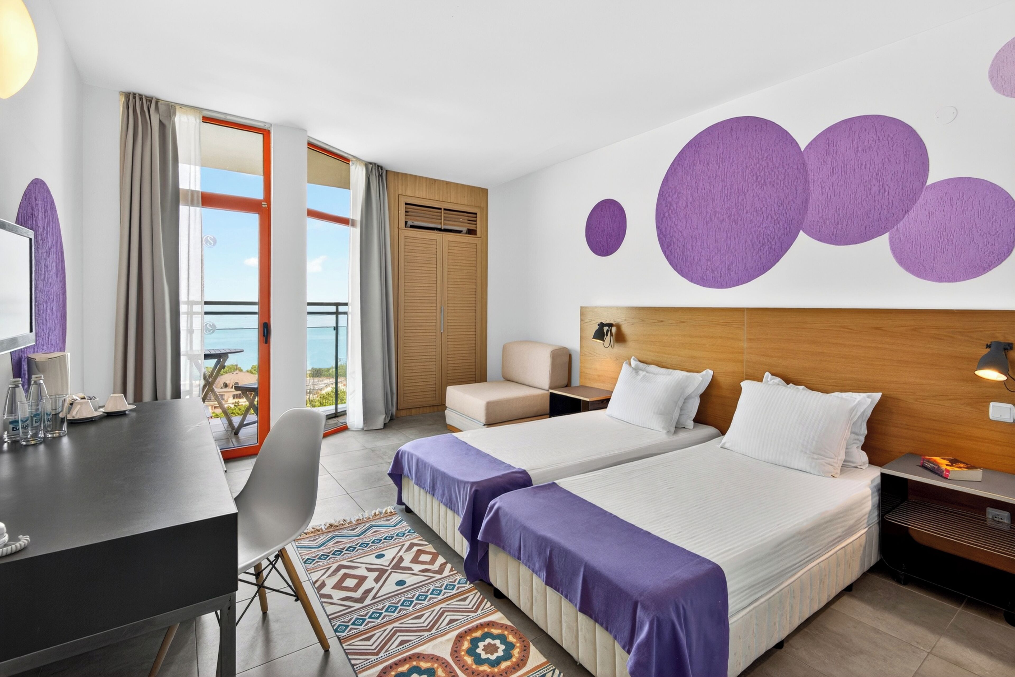A hotel room with two beds, a desk, a chair, a small table, a sofa, a patterned rug, and a view of the outdoors.