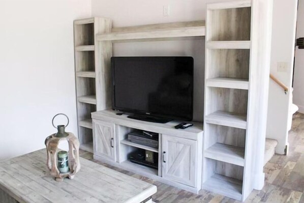 A living room with a TV on a white entertainment unit, a small table with a lantern, and a staircase to the right.