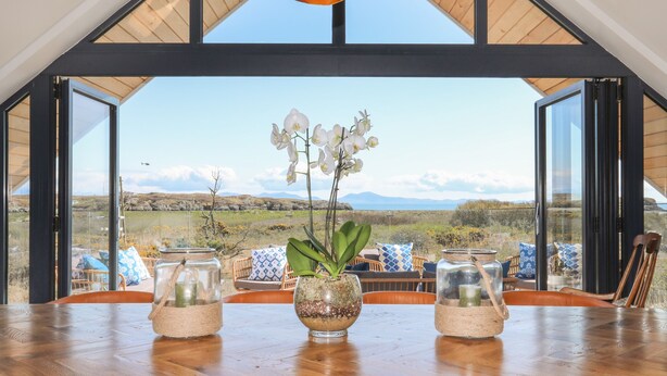 A dining table with two glass jars containing plants, a wooden table, and a view of a landscape through open doors. - Glan Towyn (Holyhead)