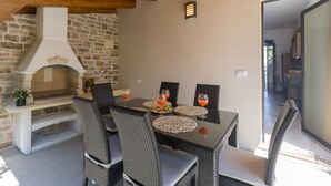 Un área de comedor al aire libre con chimenea de piedra, sillas de mimbre y una puerta de vidrio que da a un espacio interior.