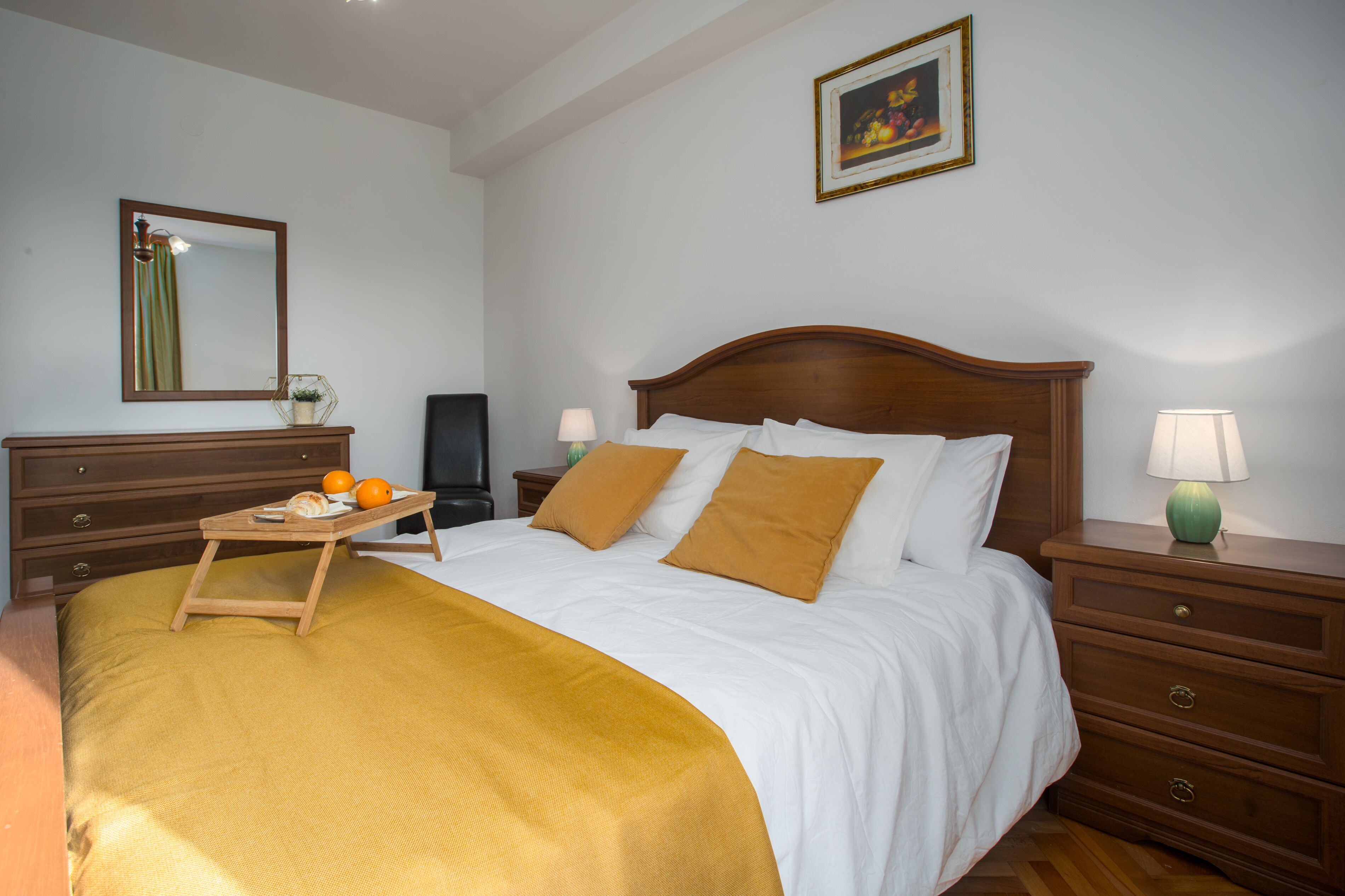 Una camera d'albergo ordinata con un letto grande, un comodino con una lampada, una cassettiera e un tavolino con arance e un vassoio.