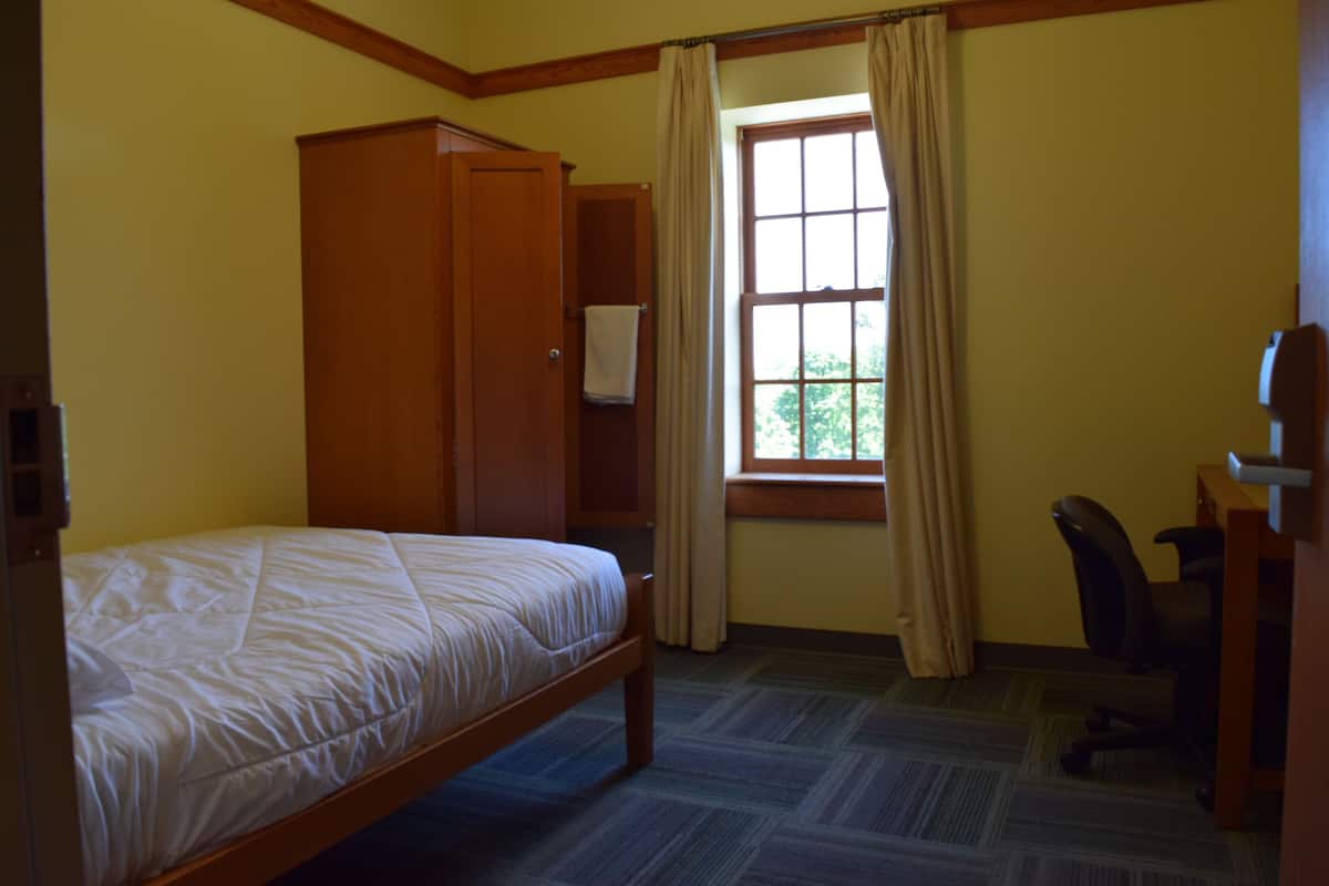 Une chambre avec un lit simple, une armoire en bois, un bureau avec une chaise et une fenêtre avec des rideaux.