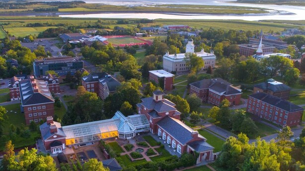 An aerial view of a campus with multiple buildings, including a large greenhouse structure, surrounded by green spaces and a body of water in the background. - Acadia University (Wolfville)