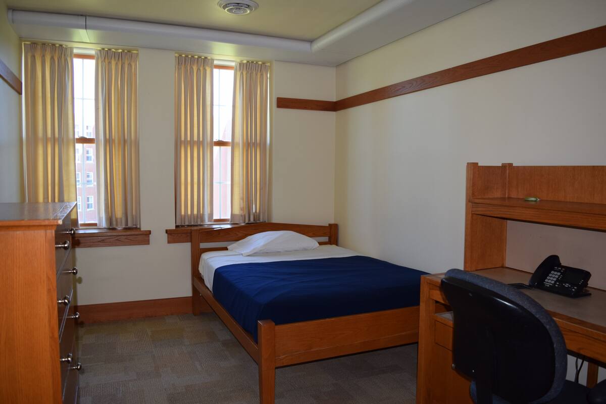 Une chambre à coucher avec un lit, un bureau avec un téléphone, une chaise et une fenêtre avec des rideaux.