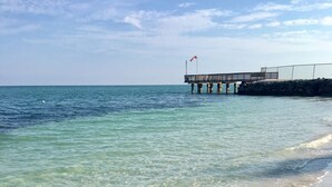 A beach with clear water, a sandy shore, and a pier with a flag. - Nice and cozy home is just a short walk to the beach and pool! (Key Colony Beach)