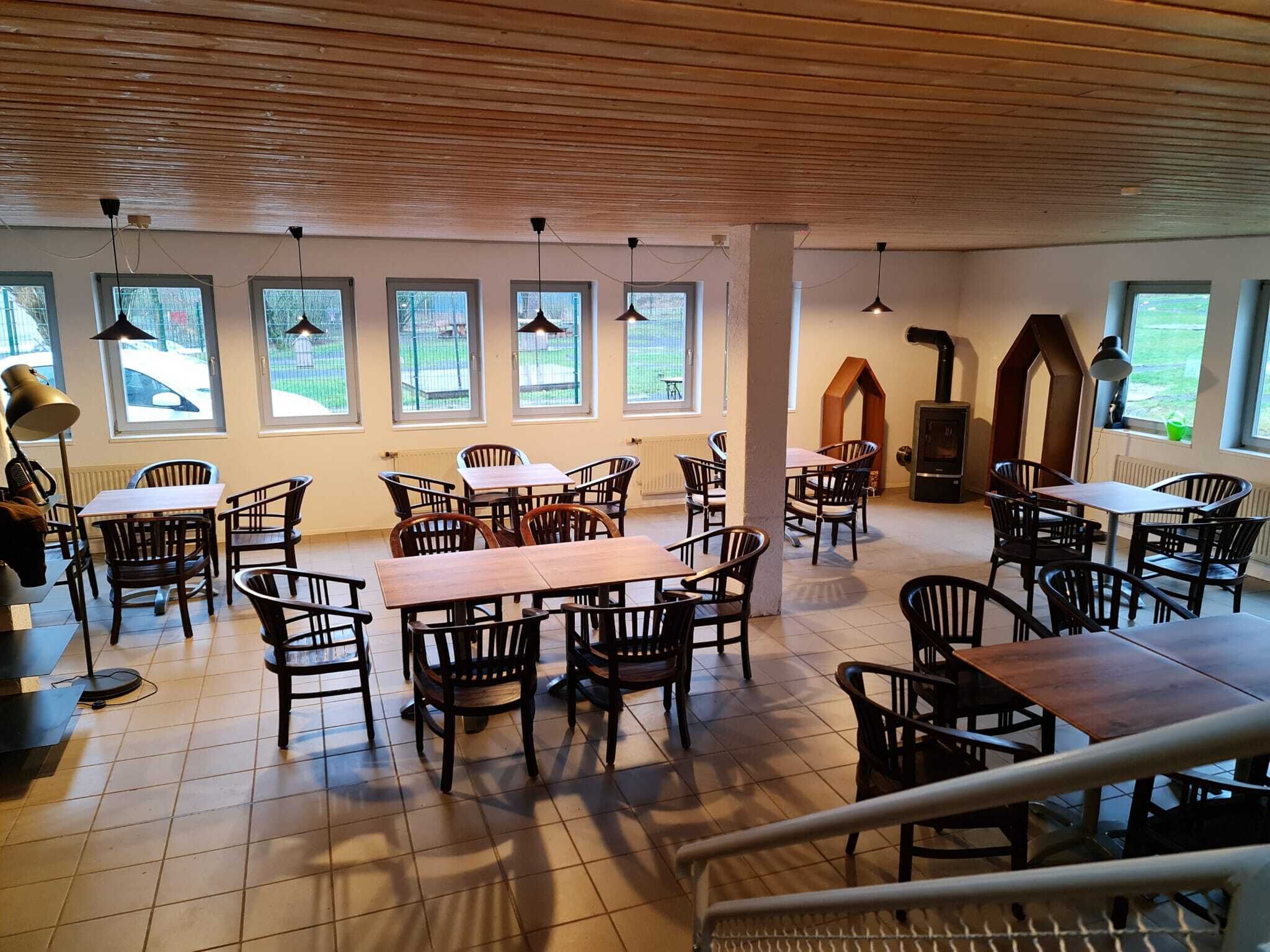 A spacious indoor dining area with multiple tables and chairs, large windows, and a wooden ceiling.