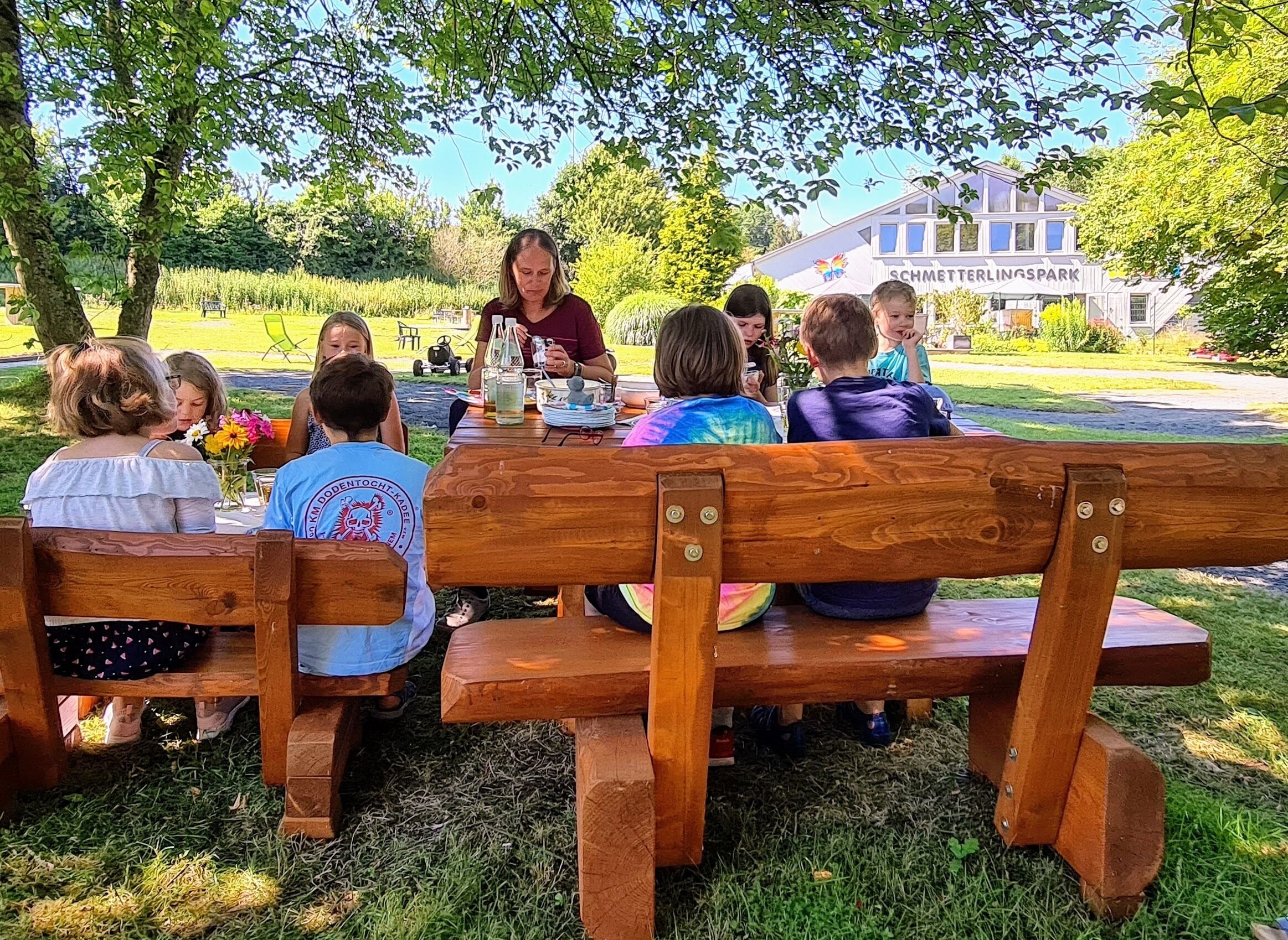 A group of people seated at a wooden table outdoors with a house in the background.