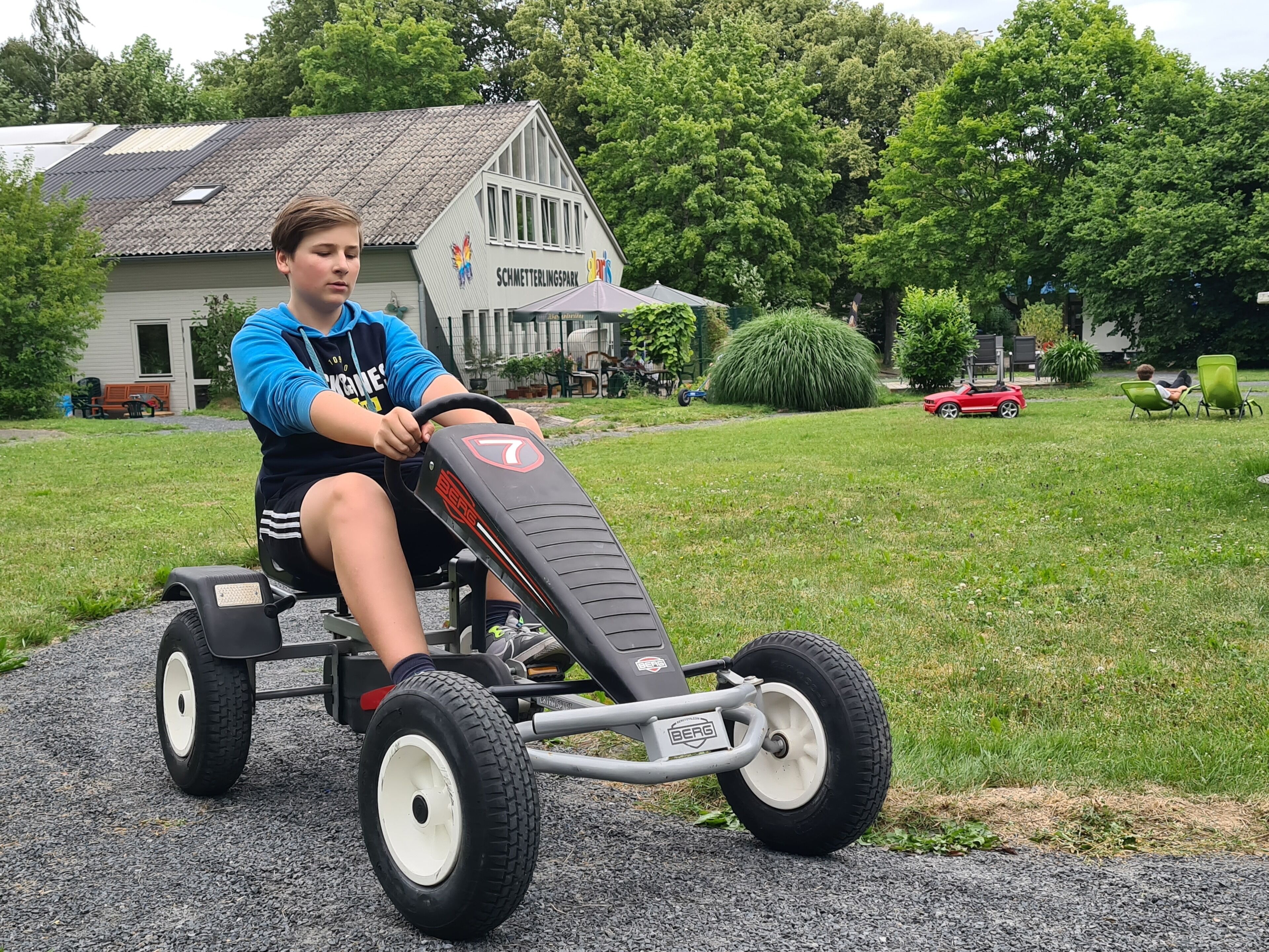 Un ni ño conduce un karting sobre un camino pavimentado, con una casa y follaje en el fondo.