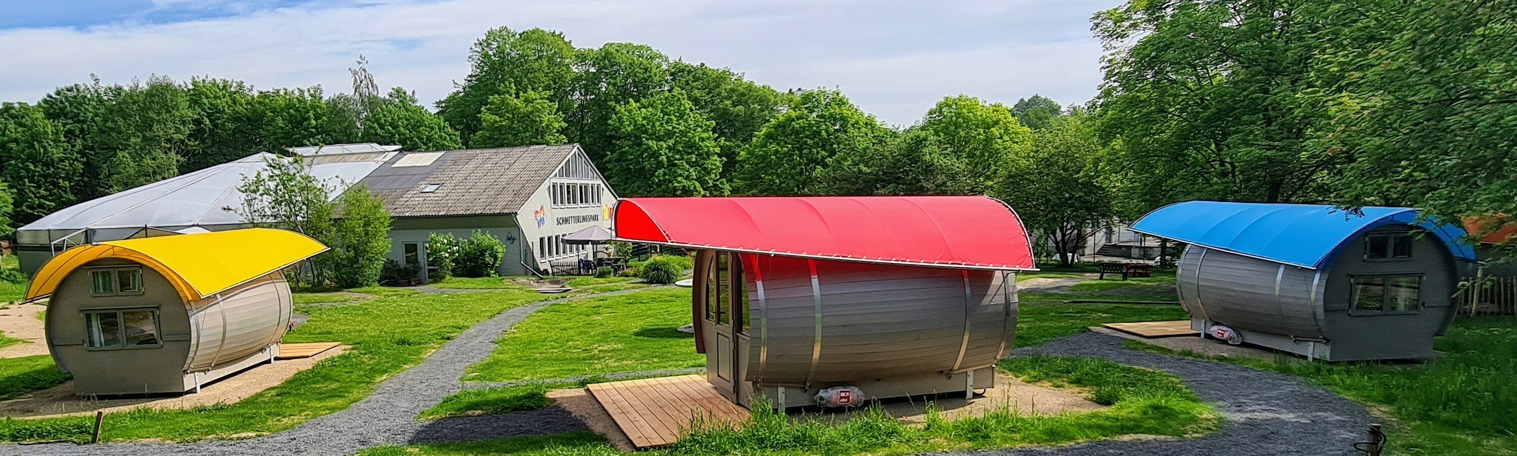 Drie kleine, kleurrijke, gelijkvloerse hutten met puntdaken, gelegen op een gazon met een pad dat naar een groter gebouw in de verte leidt.