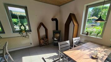 A cozy room with a wooden table and chairs, a built-in wood stove, and large windows with a view of greenery.