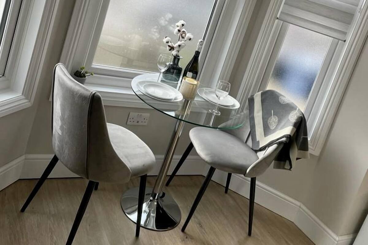 A corner dining area with a glass table, two chairs, and a view through a window with blinds.