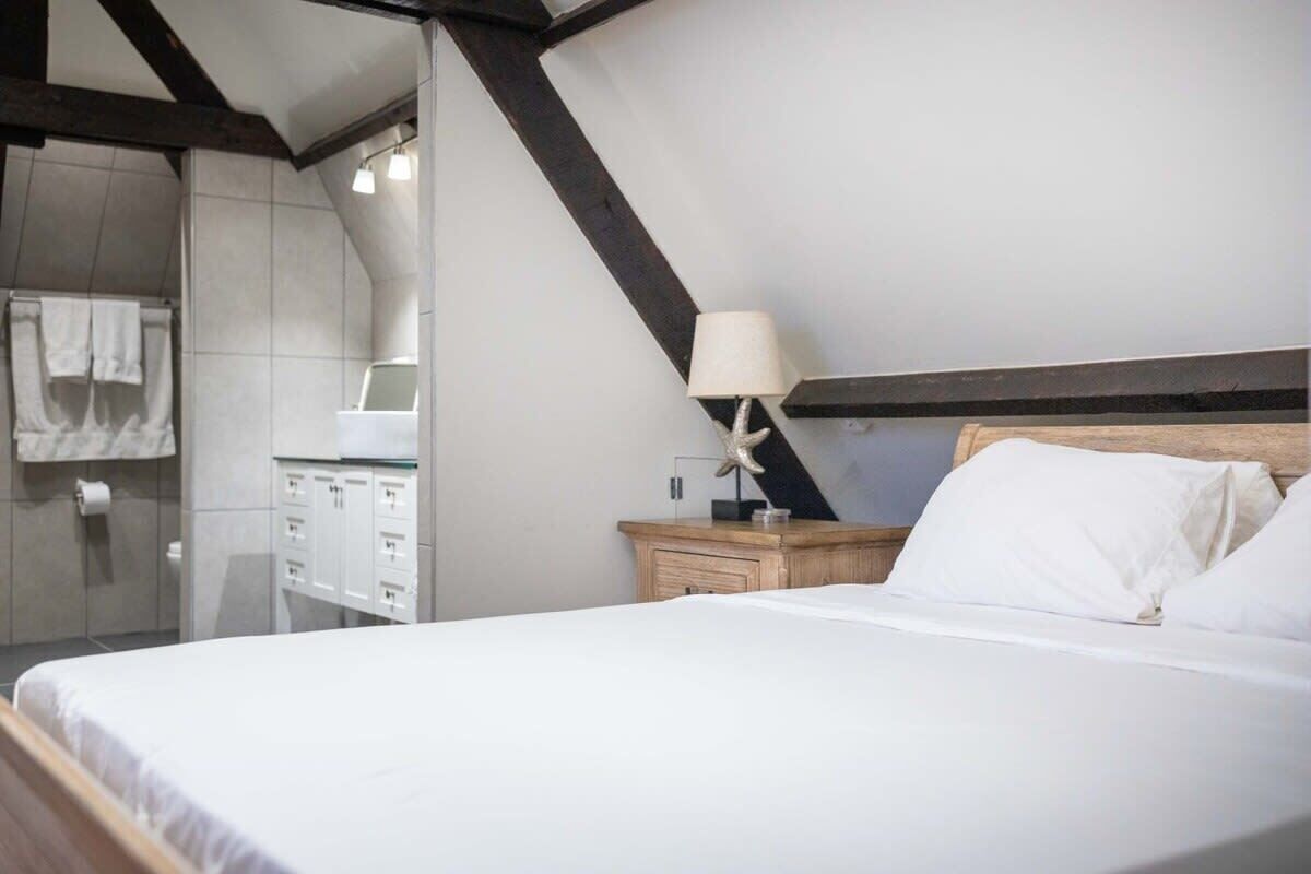 A bedroom with a large bed, a wooden headboard, a nightstand with a lamp, a wooden dresser, and a bathroom with a shower and towel rack.