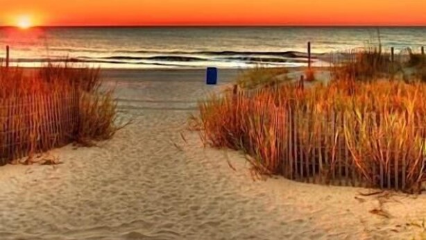 Ein Strandweg führt zum Meer, im Hintergrund ein Sonnenuntergang.