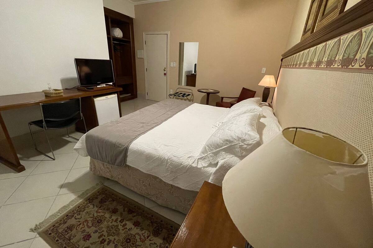A hotel room with a bed, a desk with a computer, a chair, a lamp, and a small table.