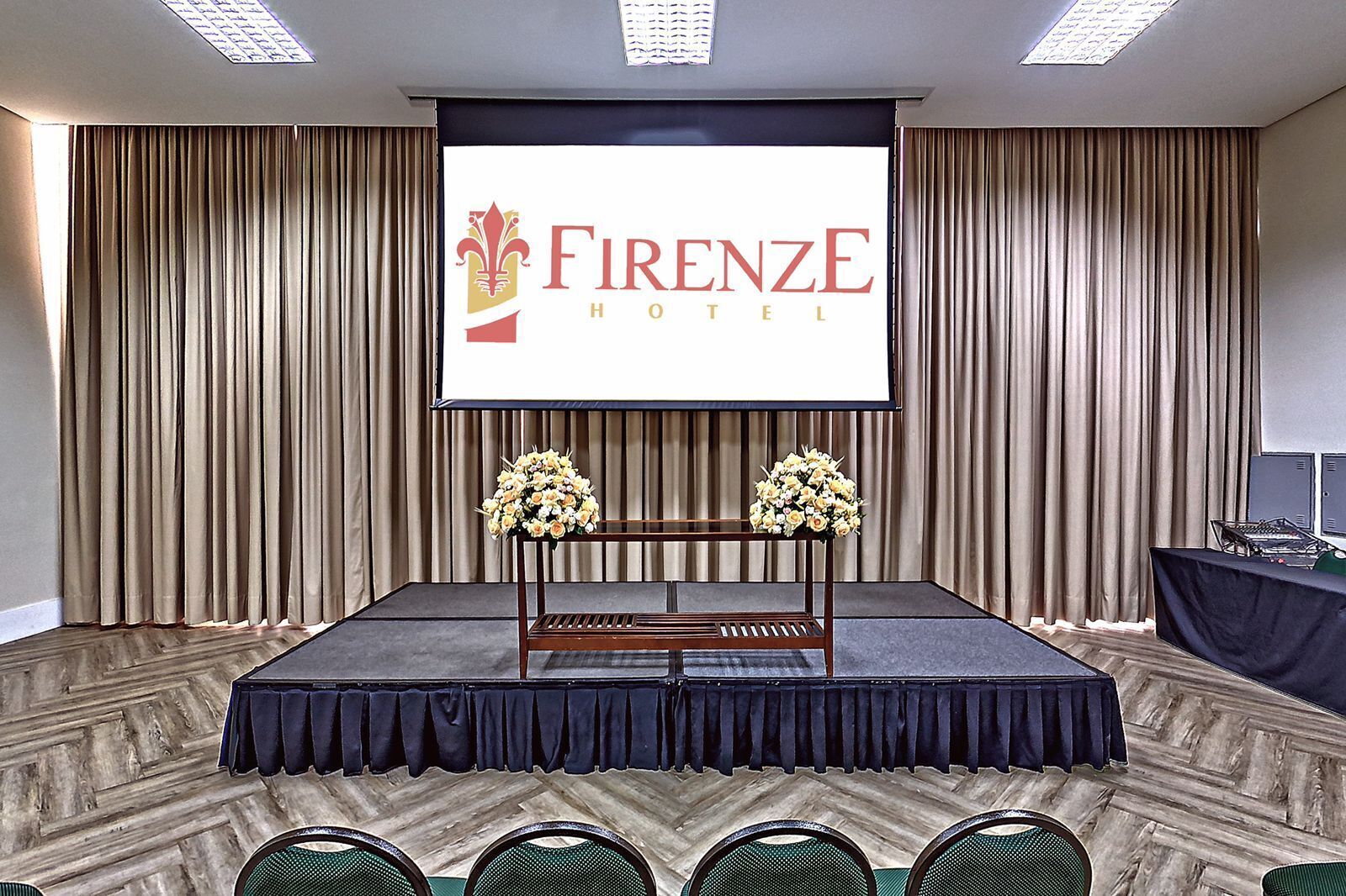 A conference room at Firenze Hotel with a large screen displaying the hotel name, a wooden podium, two flower arrangements, and a black table with chairs.