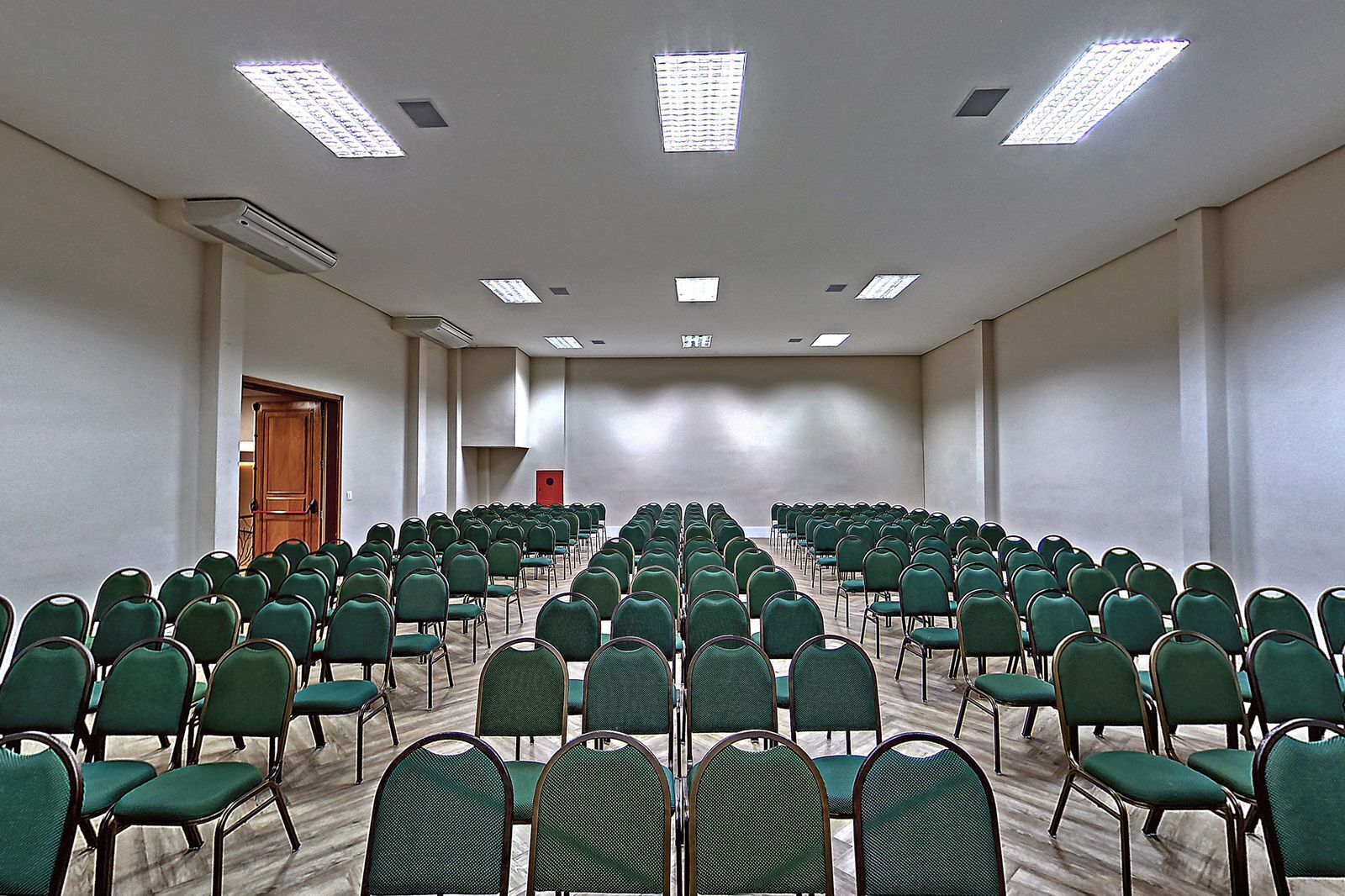 A conference room with rows of green chairs, a wooden door, and ceiling lights.