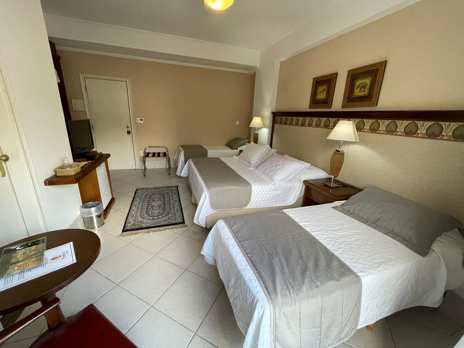 A hotel room with three beds, a desk, a chair, a small table, a trash bin, and a wall-mounted TV.