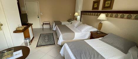 A hotel room with three beds, a desk, a chair, a small table, a trash bin, and a wall-mounted TV.