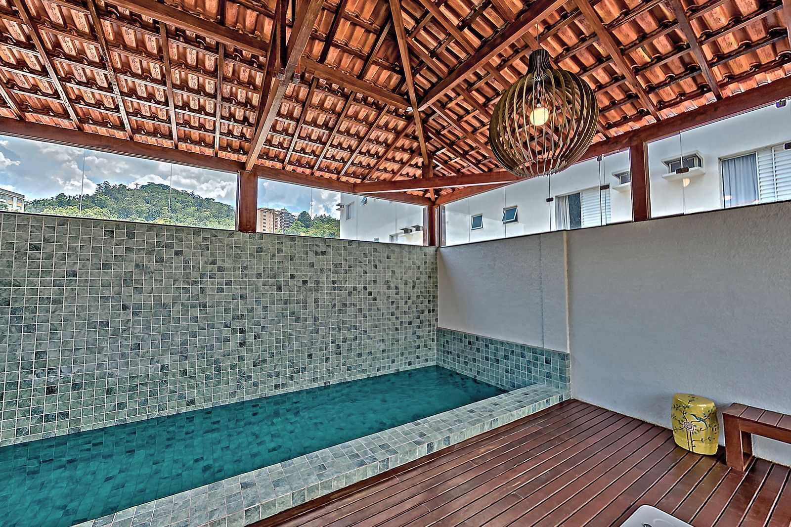 A covered swimming pool with a tiled wall and wooden deck.