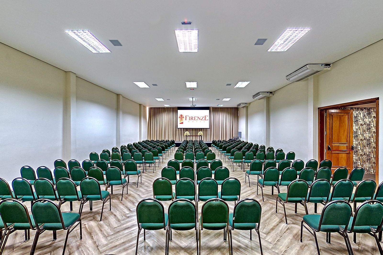 Une salle de conférence avec des rangées de chaises vertes, une scène dotée d’un écran et une porte en bois ornée d’un motif décoratif.