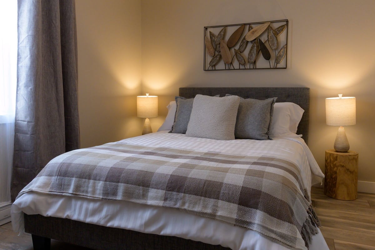 A neatly made bed with a checkered blanket, two bedside lamps, and a wall-mounted abstract art piece.