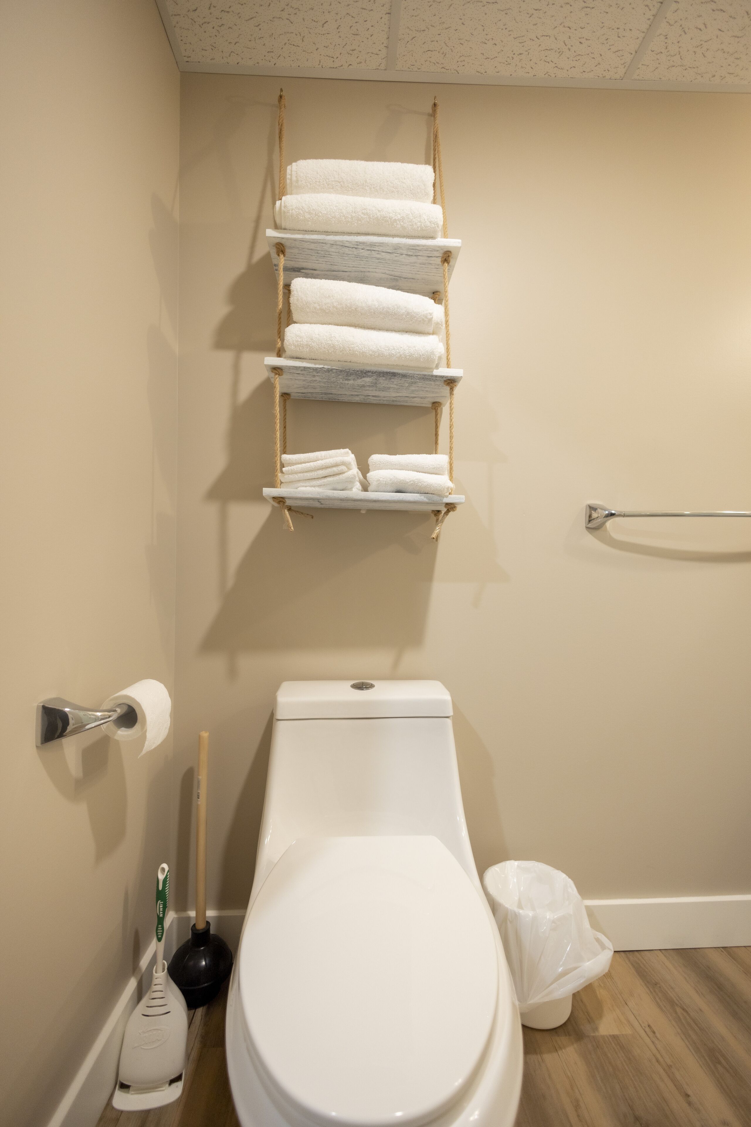 A bathroom with a toilet, a hand dryer, a plunger, a trash bin, and a towel rack with folded towels.