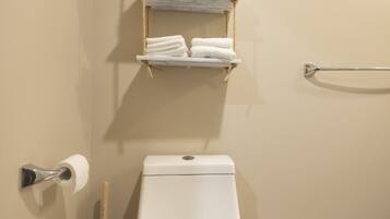 A bathroom with a toilet, a hand dryer, a plunger, a trash bin, and a towel rack with folded towels.