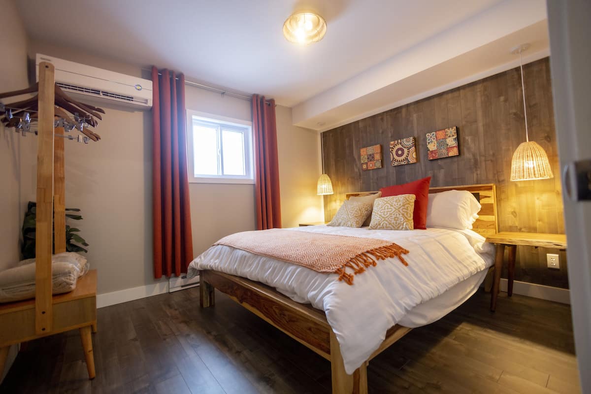 A bedroom with a wooden bed, a wooden bench, a wall-mounted air conditioner, a window with red curtains, and a wooden wall with decorative tiles.
