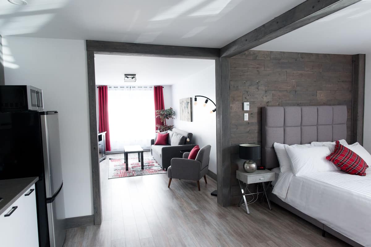 A modern hotel room with a grey upholstered bed, a small bedside table with a lamp, a grey sofa with red cushions, a red-framed window, and a wooden accent wall.