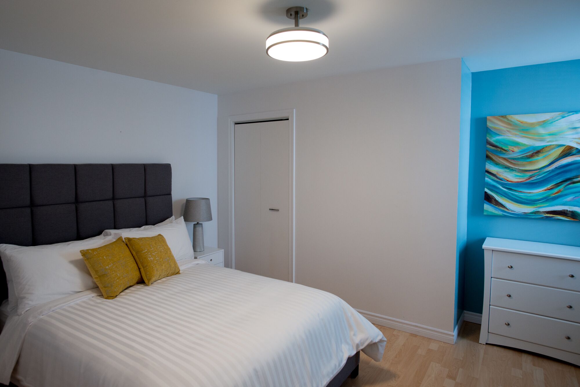 Un dormitorio con cama, mesita de noche, lámpara, puerta y pared azul con un cuadro.