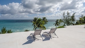 En loungestol ved stranden med to liggestole, der vender ud mod havet, palmer og frit udsyn til havet.