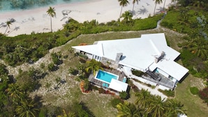 Ovenfra ses et moderne hus ved stranden med en swimmingpool, omgivet af palmer.
