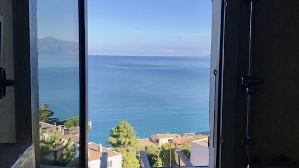 A room with a large window offering a view of the sea and a distant mountain range. - Large apartment with sea/village view 200m from the beach (Saint-Florent)