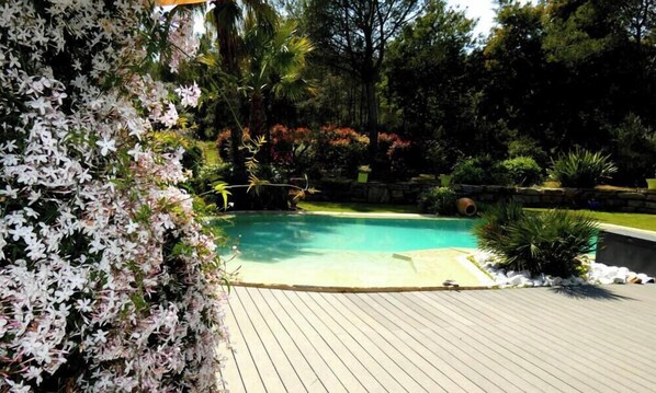 Una piscina rodeada por una terraza de madera, con un jardín que presenta flores blancas y diversas plantas.
