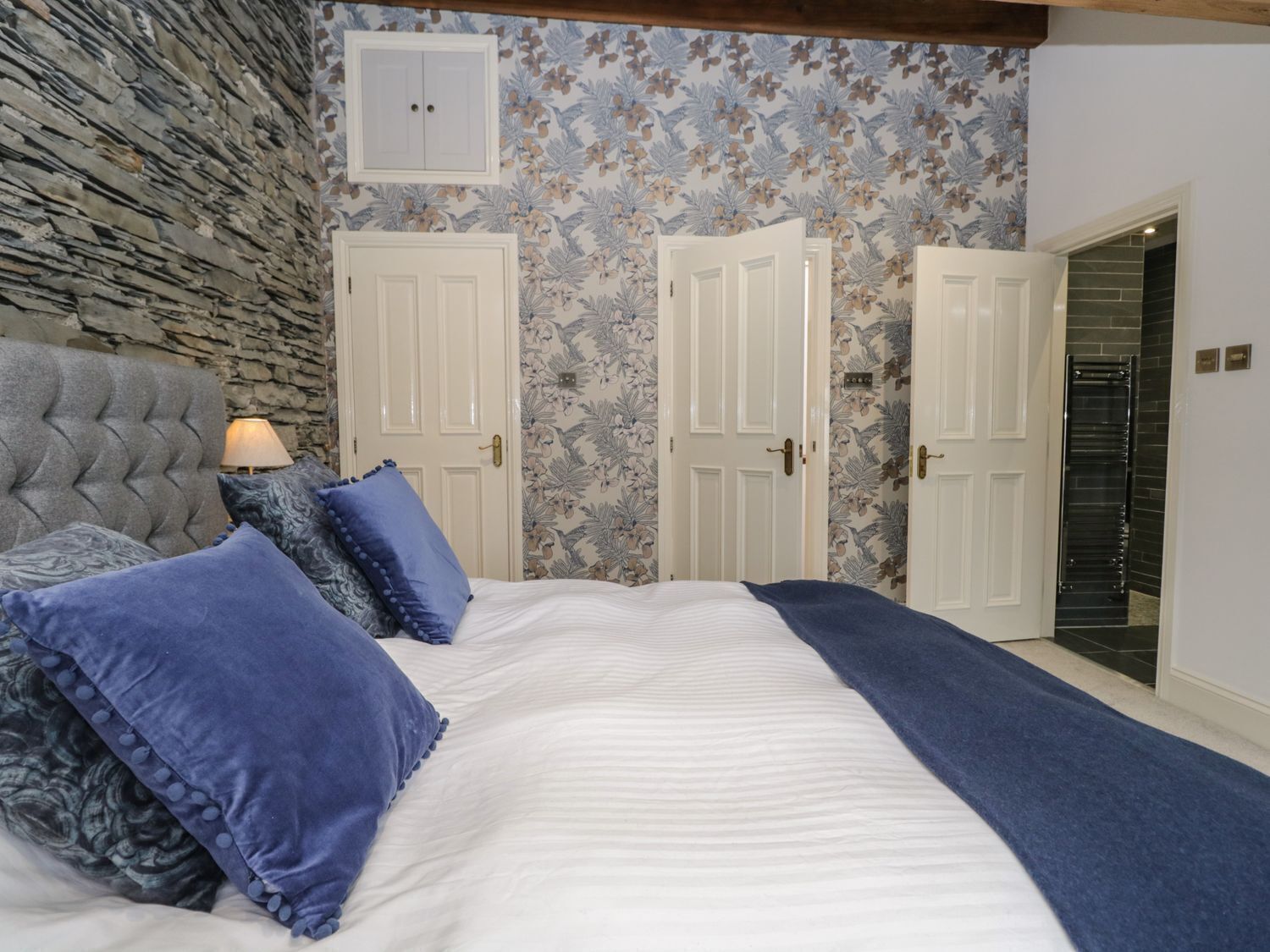 Un dormitorio con pared de piedra, dos puertas blancas, una cama con ropa de cama azul y una lámpara en una mesita de noche.