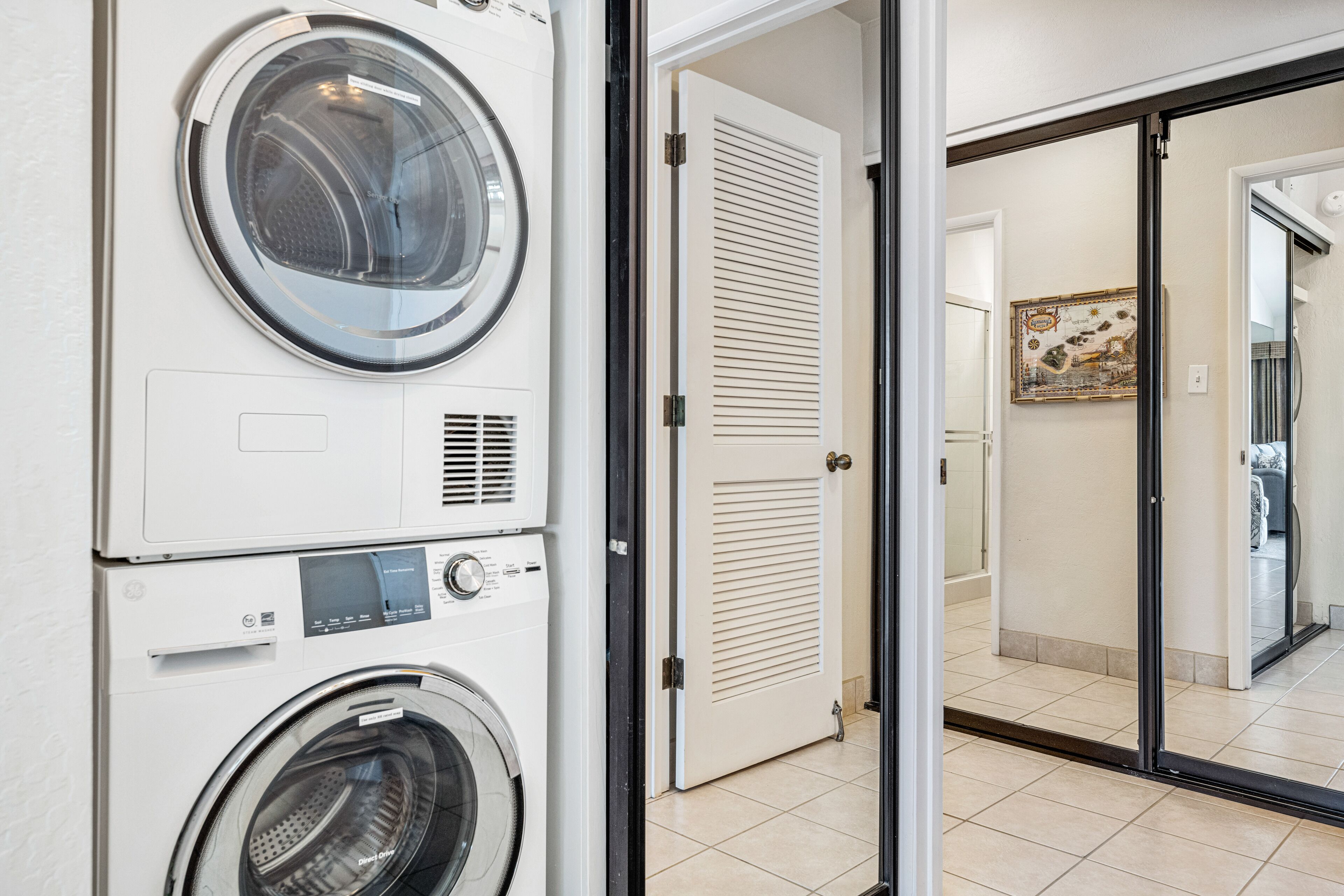 A laundry room with stacked washing machines and dryers, a door leading to another room, and a tiled floor.