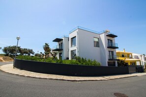 A modern two-story building with balconies, a paved driveway, and a landscaped area with shrubs.