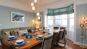A dining area with a wooden table set for six, surrounded by leather chairs. There are hanging pendant lights above the table, and a window with a view of the street is visible. - Seaside House (Weymouth)