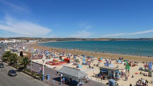 A beach with a road in the foreground, a sandy beach area with umbrellas and people, and a clear blue sky. - Seaside House (Weymouth)