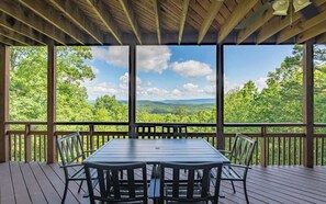 Une terrasse extérieure couverte, dotée d’une table et de chaises, de rampes en bois et offrant une vue sur un paysage boisé.