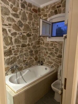Et badeværelse med stenvæg, badekar, toilet og et vindue over toilettet.
