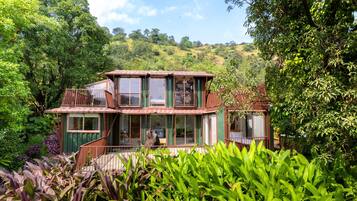 A two-story house with a balcony, surrounded by lush greenery and hills in the background.