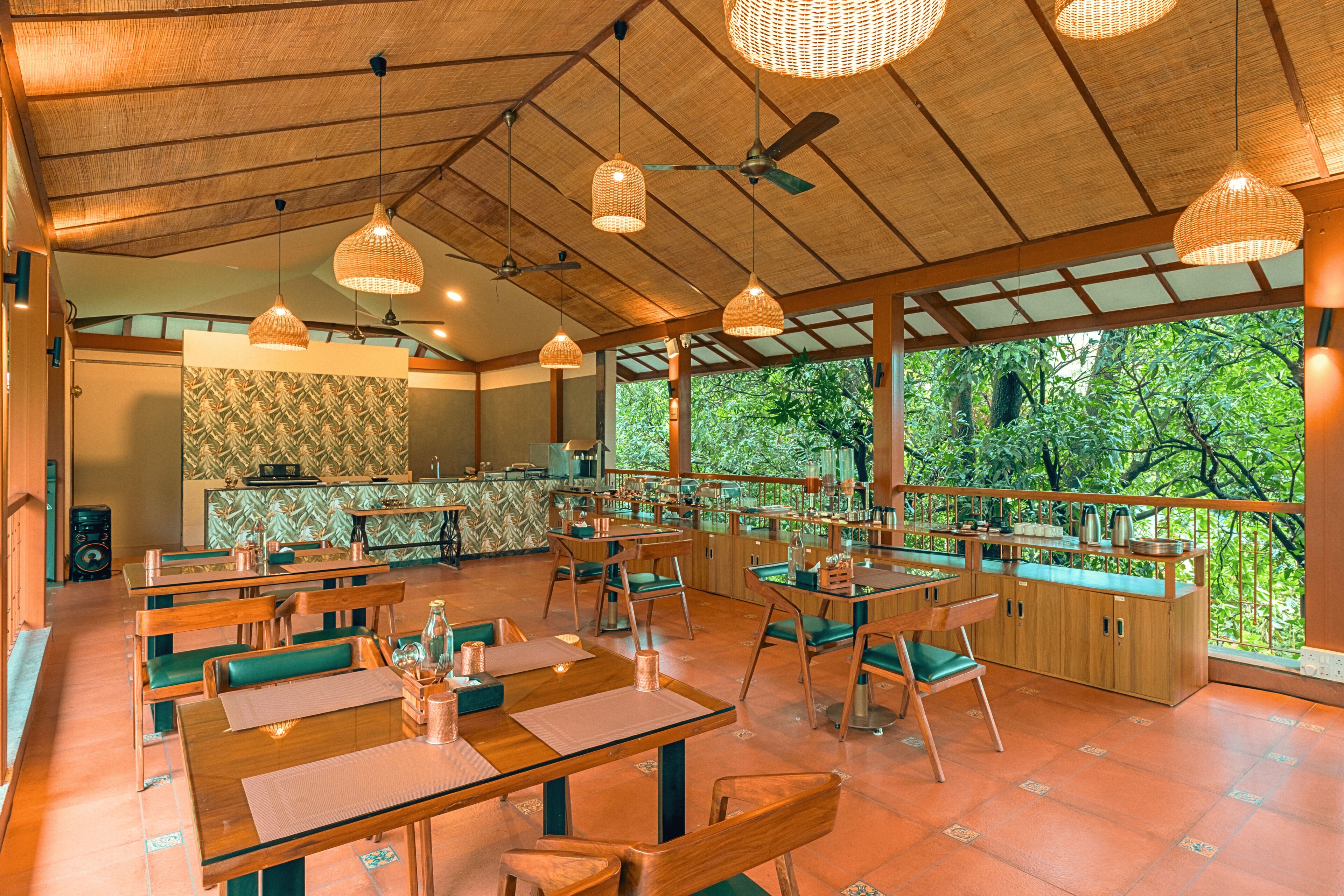 A restaurant with wooden tables and chairs, hanging wicker lamps, and a view of lush greenery through large windows.