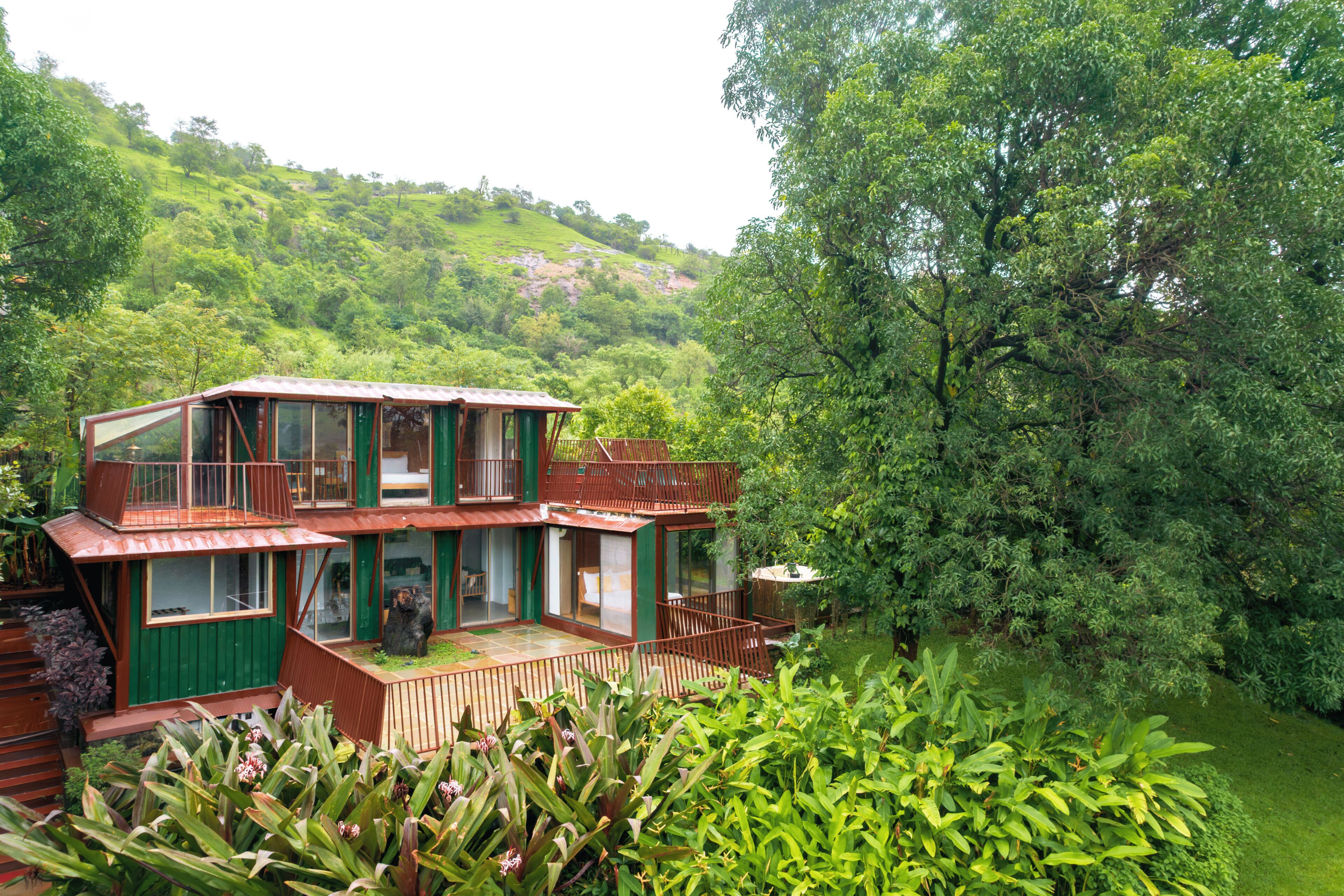 A two-story house with a balcony, surrounded by lush greenery and hills in the background.
