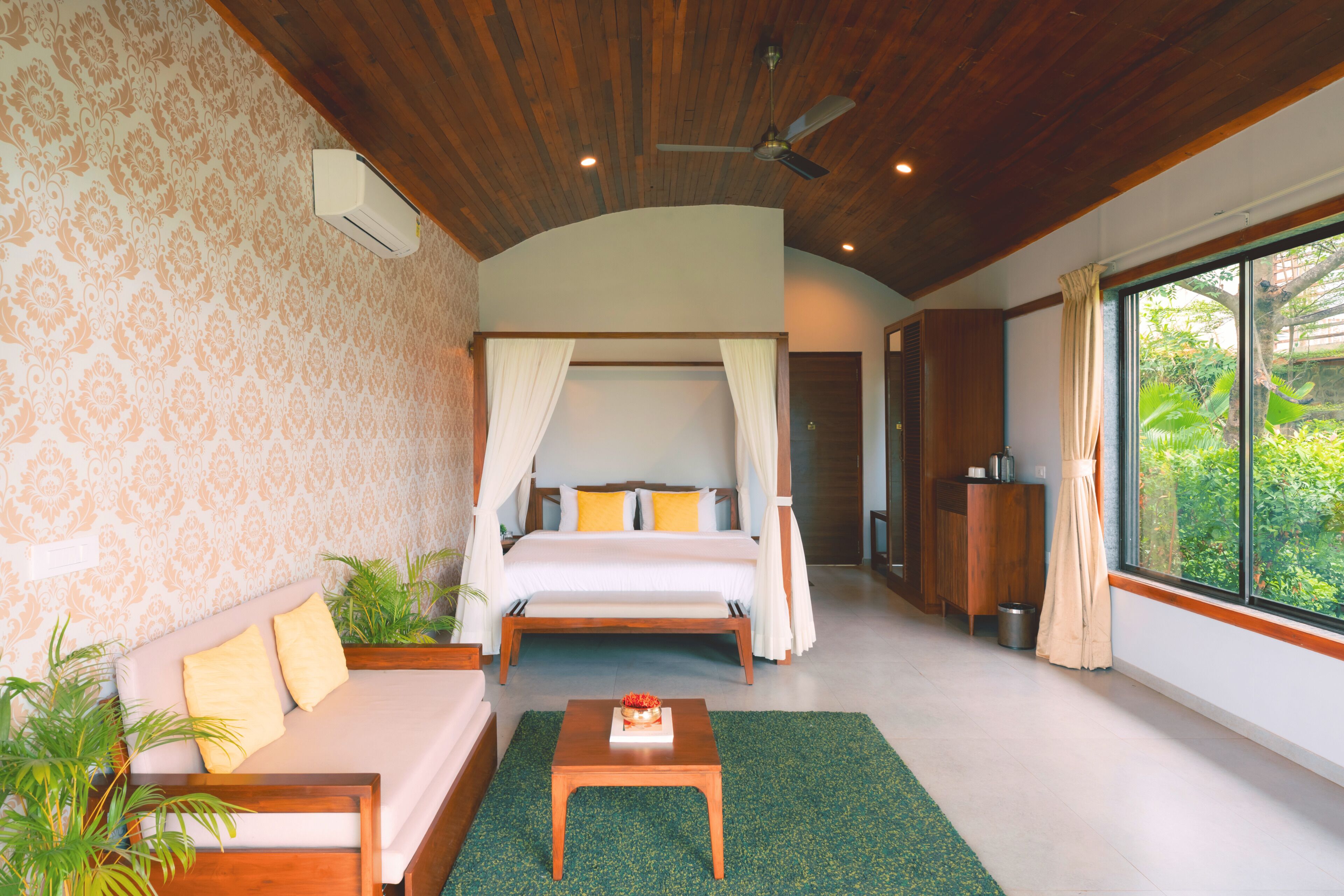 A bedroom with a canopy bed, a sofa, a small table, and a view of greenery outside.