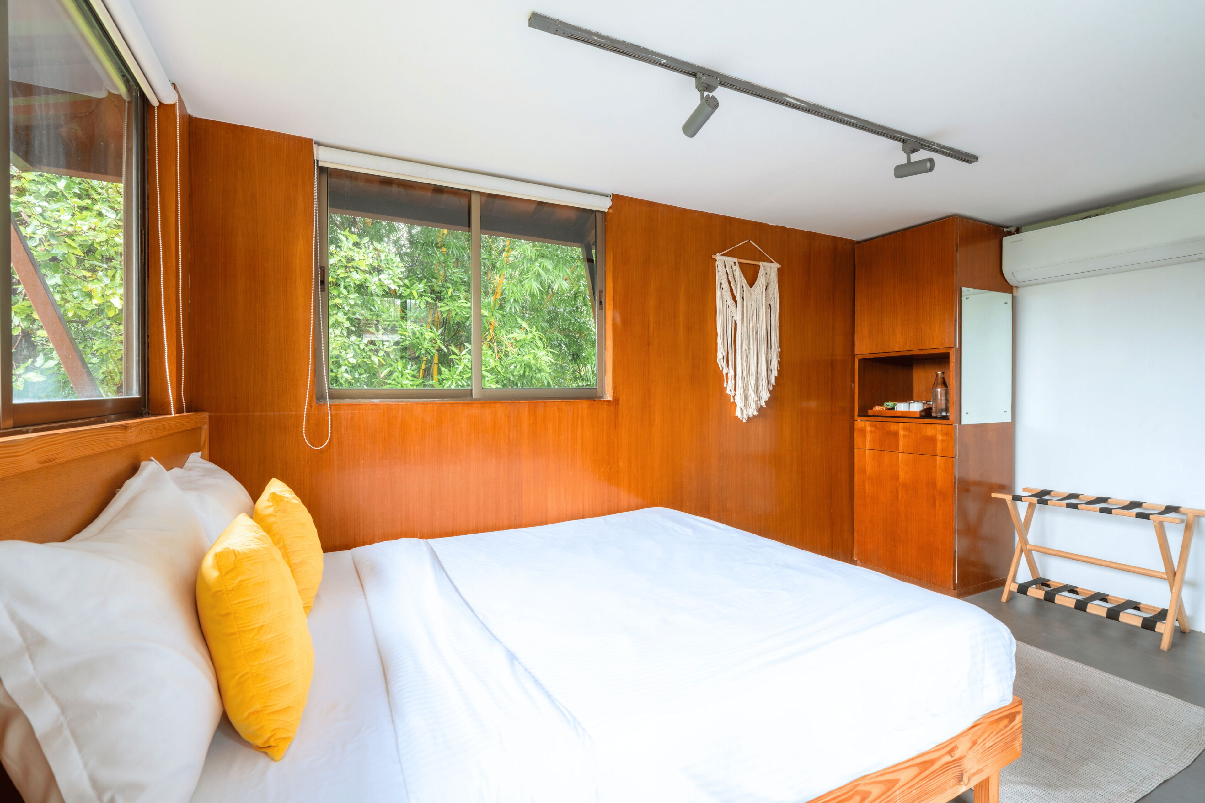 A bedroom with a wooden bed, a window with a view of greenery, a macrame wall hanging, and a small wooden shelf.